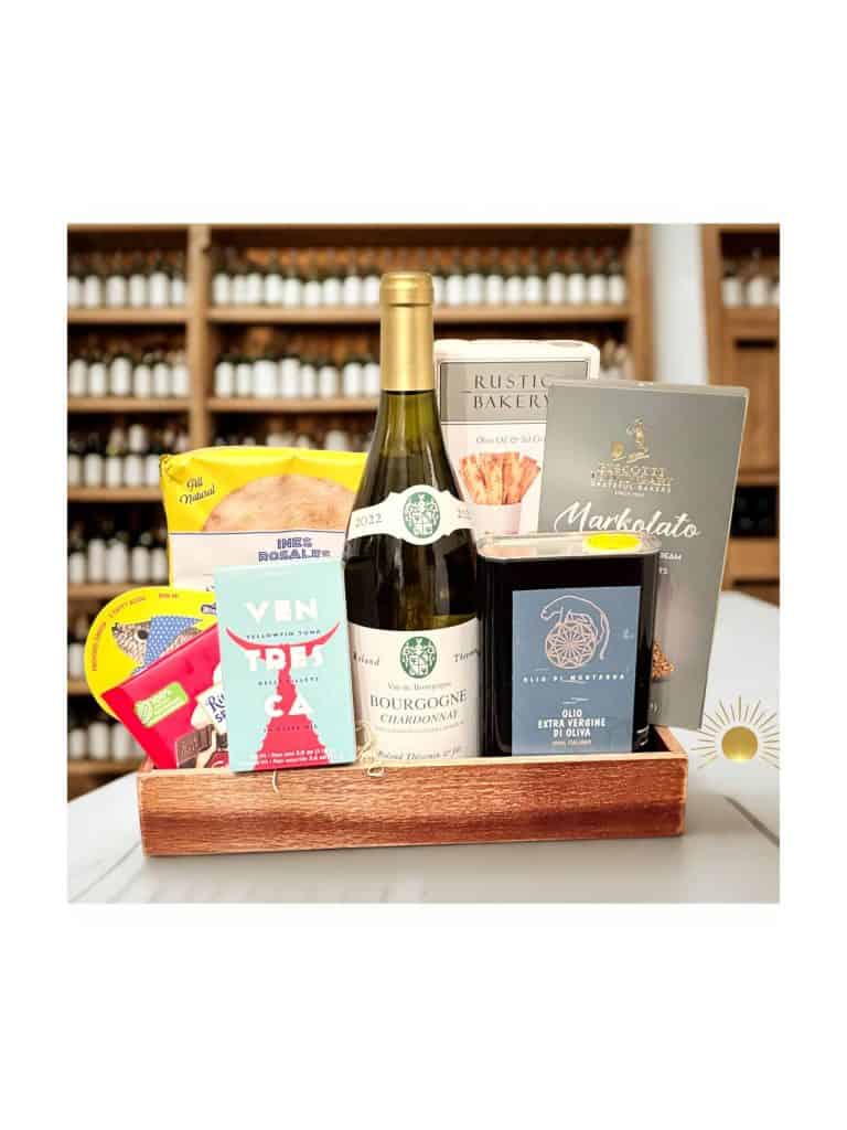 Gift hamper with a wine bottle and assorted snacks on a wooden tray in a shop setting with shelves in the background
